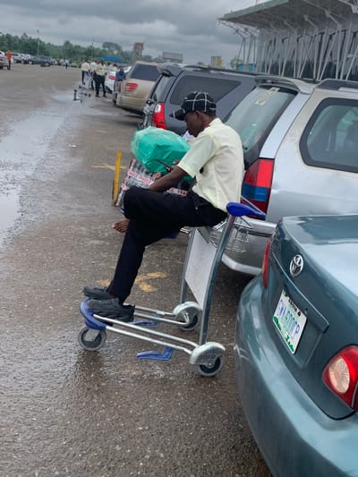 A man sits on a luggage cart with his feet on the bottom rack. He is wearing black pants, a pale yellow shirt, and a black and white checkered cap. He holds a green wrapped package and a phone in his hands. The luggage cart is silver and has four wheels. The man is positioned in a parking lot filled with cars. The ground is wet and reflects the overcast sky. In the background, several other people are visible near other luggage carts and cars. A building with a distinctive white, angled roof structure is visible in the distance. The license plate on the rear of the teal car in the foreground reads "ABUJA GMA 600CP". The scene suggests an airport or transportation hub setting.