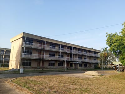 The image depicts a three-story, rectangular building with a flat roof in Tawpo, Nigeria. The building has a beige exterior with white railings on the balconies. There are numerous windows and doors visible on each floor. A street light stands in front of the building. In the foreground, there is a grassy area and a pile of sand. A parking area with a few vehicles is visible to the right of the building, under a metal structure. The sky is clear and blue. No people are visible.