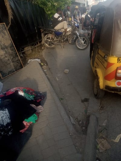 The image depicts a street scene in Mtopanga ward, Kenya. In the immediate foreground on the left, a vibrant pile of folded clothing items is displayed on a dark mat on a paved sidewalk. A large, dark rectangular board with a wooden frame leans against a black fence or barrier to the left of the clothing. Above this barrier, green foliage is visible.
On the asphalt road, a male individual wearing a black and white striped short-sleeved shirt and light-colored trousers is seated on a blue and silver motorcycle, facing right. His right hand is near his chin. Another motorcycle is partially visible behind the first. To the right of the motorcycles, a yellow three-wheeled auto-rickshaw (tuk-tuk) with "Taxi 3 Pass" written on its side is parked, facing away from the camera. It has red and white diagonal reflective tape on its rear and a license plate beginning with "K".
In the mid-ground and background, several indistinct human figures are visible on the street, some appearing to be walking or standing near what might be stalls or shops. The road surface shows signs of wear and contains scattered debris and litter. The scene suggests an active urban or commercial area.