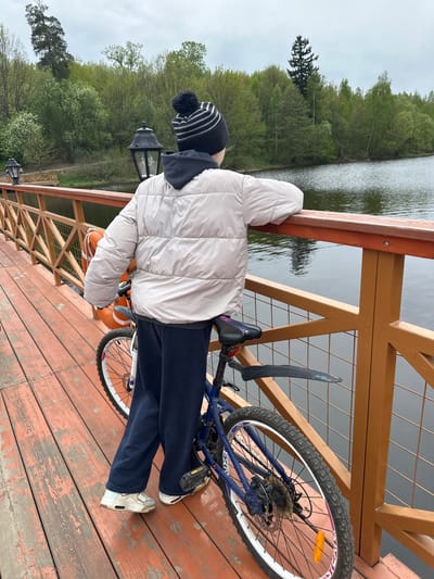 A person stands on a wooden platform, leaning against a wooden railing, with a bicycle beside them. The platform appears to be a pier or walkway extending over a body of water. The person is wearing a light-colored puffer jacket, dark pants, and white sneakers, with a striped hat. The bicycle is blue and white. In the background is a lake or river surrounded by trees. Street lamps are visible along the railing. The location is likely in the city of Grebnevo, Russia.