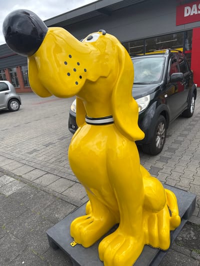 A bright yellow, cartoonish dog statue sits on a gray platform in front of a building with a red sign that reads "DAS". A black SUV is parked behind the statue, and a silver car is visible to the left. The ground is paved with gray rectangular tiles. The sky is overcast. The statue is in Gronau, Germany, at the location of a former "DAS"-branded electronics store.