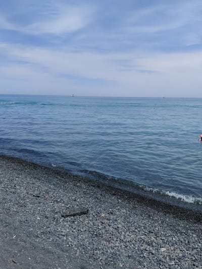 A wide shot captures a pebble beach in Ақши, Kazakhstan, meeting the calm, blue waters of a large body of water. The sky above is a pale blue with scattered, wispy white clouds. The beach is composed of numerous small, gray pebbles, with a few larger, darker stones and a patch of asphalt near the foreground. The water exhibits gentle ripples and a slight wave breaks at the shoreline. In the distance, two small boats are visible on the water, and a person can be seen partially submerged in the water on the right side of the frame. A few orange buoys are also dotted across the surface of the water.