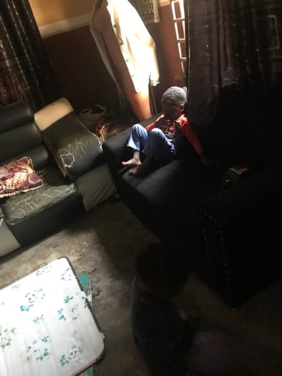 This image depicts an interior scene in Oba-Ile, Nigeria, possibly a living area, characterized by dim lighting and a worn appearance. The background wall is dark reddish-brown. A window on the right side, partially obscured by dark, patterned curtains, allows natural light into the room. A light-colored garment, possibly a jacket with green accents, hangs on a stand or hook against the back wall.
In the center-right, a young male child, barefoot, sits on a dark-colored sofa. He wears a long-sleeved shirt, red on top and dark below, paired with blue trousers. His left hand is near his mouth or chin, and his gaze is directed downwards. A small electronic device with red buttons rests on the right armrest of this sofa. To the left, a section of a black faux leather sofa shows significant wear, with cracked and peeling upholstery revealing a lighter material underneath. A patterned cushion, featuring brown, red, and beige hues, rests on this sofa. The floor appears to be a concrete or hard surface, showing some dirt or debris.
In the lower foreground, partially obscured and heavily shadowed, another young child is visible, possibly sitting on the floor or a low surface. Only their head and upper body, dressed in dark attire, are discernible. A low table or surface, covered with a white cloth featuring a light green floral pattern, is positioned in the lower-left foreground, partially out of frame.