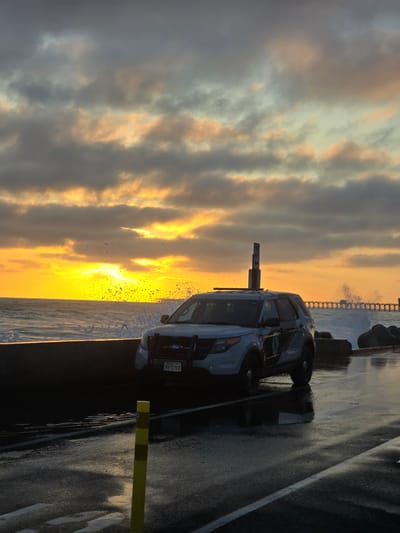 A police SUV is parked on a wet asphalt road overlooking the ocean at sunset in Oceanside, California. The sky is filled with dramatic clouds painted in shades of orange and yellow from the setting sun, which illuminates the crashing waves and sea spray. The car, a white Ford Explorer, is positioned on the left side of the frame, facing forward. Its emergency lights are not visible, and it appears to be unoccupied. A yellow and black striped bollard stands in the foreground on the left. In the background, a long pier stretches out into the ocean, with waves breaking around its pilings. The road surface is wet and reflective, suggesting recent rain or sea spray. The overall scene depicts a moody and atmospheric coastal setting at dusk.