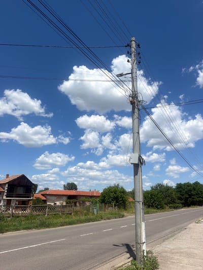 This image depicts a roadside scene in Plodovitovo, Bulgaria. The foreground features a paved road with a white dashed line marking the center. To the left of the road, a concrete utility pole stands tall, adorned with numerous electrical wires, a street lamp, and a gray electrical box. Behind the pole, a weathered fence with an ornate pattern separates the road from a residential area. Several houses with red tile roofs are visible, interspersed with lush green trees and vegetation. The sky above is a vibrant blue, dotted with fluffy white cumulus clouds, suggesting fair weather. The overall impression is one of a quiet, rural setting on a sunny day. There are no people or animals present in the image.