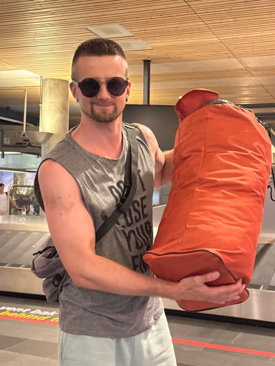 The media file shows a man holding a large orange duffel bag. He is wearing a grey tank top, light blue shorts, and round, dark sunglasses. The man has a beard and short hair. He appears to be smiling.

The background suggests an indoor setting, possibly an airport or a train station, with wooden paneling on the wall and what looks like a luggage carousel. There are also glimpses of a window showing an outdoor scene with trees and possibly mountains, indicating a travel context. The lighting suggests it could be daytime. There is no clear indication of the weather. The man's expression and the way he is holding the bag suggest a relaxed or pleased emotion.

Visible text includes "USE" and part of another word, "SE", on the man's tank top. Another partially visible text on a red and yellow sign at the bottom reads "vent bak behind".

The overall scene depicts a man with his luggage, likely at a transportation hub, preparing for or having just completed a journey.