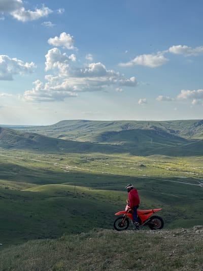 A view of a motocross bike on a grass-covered hill, possibly in the Орджоникидзевский поселковый совет region of Ukraine. The motorcycle, predominantly orange in color, is oriented towards the viewer's right, with a rider dressed in dark pants and a red jacket. The rider is wearing a helmet. The backdrop features rolling green hills and mountains. The sky, visible on the left side of the image, is a bright blue with white, puffy clouds.