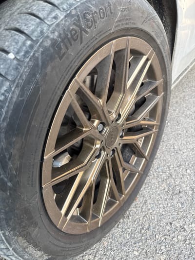 The image shows a close-up of a bronze-colored, multi-spoke alloy wheel with a tire mounted on it. The wheel is resting on an asphalt surface. The tire has text on its sidewall, including "TRIANGLE GROUP", "MADE IN CHINA", "ZEEX", and "OUTSIDE". A silver valve stem cap is visible on the wheel. The scene appears to be outdoors, possibly in a parking lot or roadside, given the asphalt and the slight blur suggesting movement or a tilted perspective. The lighting appears to be natural, suggesting daytime. There are no people visible in the image. The overall impression is of a car wheel, likely removed or in the process of being changed, with some wear and tear visible on the tire. The location context provided is Kaspichan, Bulgaria, but there are no specific visual cues in the image to confirm this location.