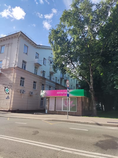 The image shows a street scene in Polatsk, Belarus. The foreground is an asphalt road with double white lines. A sidewalk runs alongside the road, with a curb separating them. Behind the sidewalk is a building with a storefront. The storefront has a pink and green awning with the word "КОСМЕТИК" (Cosmetic) and the logo "иос" visible. In front of the storefront is a pole with a circular traffic sign indicating a speed limit of 40. To the left of the storefront, a beige building with windows and air conditioning units is visible. Further to the left, a sign with a green cross symbol and the number 18 is hanging. A large tree with green foliage stands to the right of the storefront, partially obscuring a light blue building behind it. The sky is blue with scattered white clouds. Several small dark objects, possibly birds, are visible in the sky.