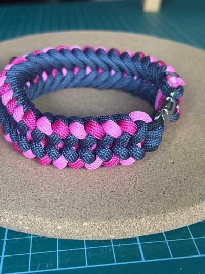 A close-up shot showcases a handcrafted paracord bracelet. The bracelet is intricately woven with two distinct colors: a dark grey and a vibrant pink. The weaving pattern creates a textured, braided appearance with the pink cords peeking through the grey in a repeating sequence. The bracelet has a metallic clasp, likely made of stainless steel, which is partially visible at the top of the frame. The bracelet is resting on a circular, textured surface that appears to be cork or a similar material. In the background, to the left, a blurry teal surface is visible, and to the right, a portion of a green cutting mat with a blue grid pattern is present. The lighting suggests an indoor setting, possibly a craft or workspace. No people are visible, and the focus is entirely on the handmade object. The overall impression is one of detailed craftsmanship and colorful design.