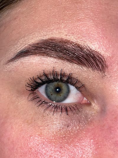 The image is a close-up, angled shot of a person's eye and eyebrow, with a focus on the skin texture. The eye is open, revealing a light blue-green iris with brown speckles. Dark mascara is applied to the eyelashes, making them appear thicker and longer. The eyebrow, located in the upper left of the frame, is dark and well-defined with visible hairs. The skin surrounding the eye and eyebrow is fair and appears to be slightly shiny, possibly due to oil or makeup. There are visible pores and fine lines on the skin. The background is out of focus, but suggests a natural skin tone. The lighting appears to be soft, highlighting the texture of the skin and the details of the eye and eyebrow. No specific time of day, weather, location cues, emotions, or visible text are evident in the image.