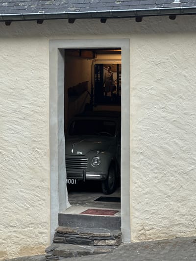 The image shows an interior of a garage or shed, viewed from an exterior doorway. A vintage gray car, possibly a Fiat 500 or similar model, is parked inside. The license plate is visible and reads "3001". To the left of the car, a small rug with a red pattern is visible on the floor. In the background, a poster or picture of a woman in a dress is displayed on the wall. The exterior of the building features a textured cream-colored wall and a dark-colored roof with visible shingles. Stone steps lead up to the doorway. The scene is located in the city of Vianden, Luxembourg.