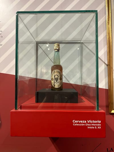 A single, clear glass display case with a green frame encloses a vintage beer bottle. The bottle is green glass with a gold foil cap and a label featuring an eagle crest and Spanish text. It rests on a black, rectangular pedestal, which sits on a red, tiered display base. The background is a wall with diagonal stripes of pink and white. Text on the red base reads "Cerveza Victoria Colección Diez Morodo Inicio S. XX". This display is located in a museum in Toluca, Mexico.