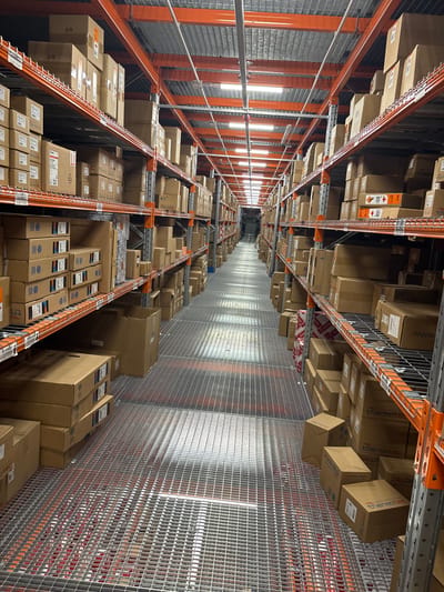 This image shows a narrow aisle in what appears to be a large storage warehouse. Tall metal shelving units, painted orange, stretch towards the vanishing point. These are packed with cardboard boxes of varying sizes, many bearing labels. The floor and walls of the aisle are made of wire mesh, creating a gridded effect. Rows of fluorescent lights are visible along the ceiling. There is no indication this image is from Gara Elin Pelin, Bulgaria.