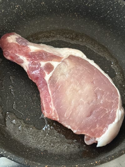 An overhead shot shows a thick, raw pork chop with a bone, lying in a black, speckled non-stick frying pan with a small amount of cooking oil. The pork chop is uncooked and displays a pale pink to reddish hue with visible marbling and a layer of white fat along the edges. The oil in the pan is clear and shows small bubbles, indicating it is heating up or has recently been heated. The background is dark and out of focus, with the focus primarily on the pork chop and the immediate surroundings within the pan. There is no indication of the location being in the city of Nogir, Russia.
