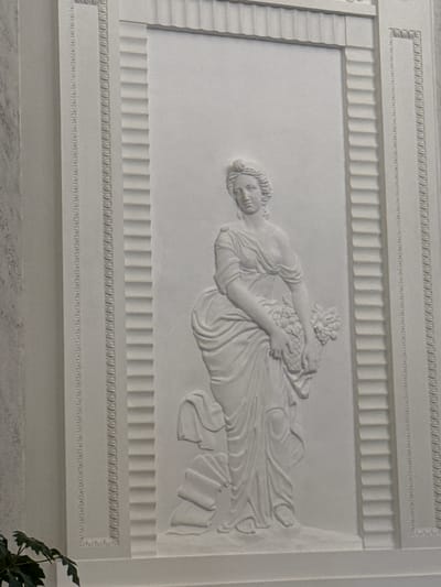 The image displays a white bas-relief sculpture on a wall, depicting a female figure in classical attire holding a basket of fruit and flowers. The sculpture is framed by decorative borders featuring geometric patterns and fluted details. The overall aesthetic is one of classical artistry and grandeur. The location of this sculpture is identified as the Imperial Stables in Strelna, Russia.