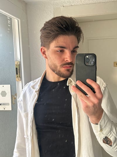 The image displays a young adult male taking a mirror selfie in an indoor setting. The male, appearing Caucasian, has dark brown, medium-length hair styled upwards and to the side, a trimmed beard, and a mustache. He is dressed in a black crew-neck t-shirt under an unbuttoned, light-colored, textured shirt with visible distressed edges on the collar and shoulders. A tattoo showing the letters "GAU" is partially visible on his left forearm. He holds a black smartphone, identifiable as an iPhone with a three-lens rear camera system, in his right hand, angled to capture his reflection. The background features a light-colored, textured wall. To the subject's left, a white door frame is visible, containing a metallic strike plate. Below this, a white intercom system with a speaker grille and a label reading "P. CAMILLO" is mounted on the wall. A partial white sign with the letter "T" is visible in the upper right quadrant of the reflection. The specific location within Rafz, Switzerland, is not discernible from the visual evidence. The scene is consistent with an interior entryway or hallway.