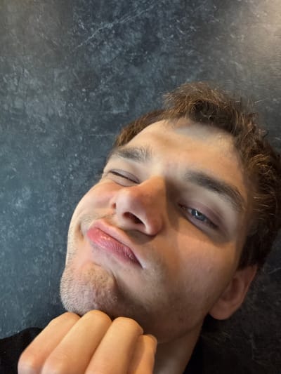 A close-up, tilted selfie captures a male individual appearing to be in his 20s or 30s, situated in Minhang District, China. The subject has dark brown, disheveled hair and short stubble visible on his chin and jawline. His right eye (viewer's left) is closed or squinted, while his left eye (viewer's right) is partially open, revealing a blue iris. His lips are prominently pursed into a pout, and his right hand (viewer's left) is curled, with fingers supporting the chin/jaw area. He wears a dark-colored garment, possibly a shirt, visible at the lower left. The background is a dark gray to bluish-gray textured surface with lighter, irregular patterns resembling marble or stone. Soft, diffused lighting illuminates the scene, casting minimal shadows.