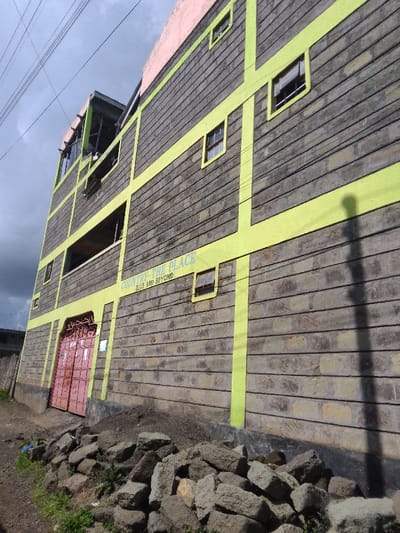 The scene depicts the exterior of a four-story building, located in Njoro ward, Kenya, viewed from a low-angle perspective. The building's facade is constructed of grey, horizontally-laid masonry blocks, intersected by distinct bright yellow-green vertical and horizontal painted bands. The uppermost visible section of the building is light pink. Multiple rectangular windows, some open, are present on various floors, accented with yellow-green frames. On the ground floor, a secured pink double metal gate serves as the primary entrance. Above this entrance, on the second floor, the text "COUNTRY-THE PLACE" followed by "2019 AND BEYOND" is painted in yellow-green. The third floor exhibits a wide, open-air section or balcony with a visible railing, containing indistinct white objects. The ground in the foreground consists of dirt and scattered green vegetation, with a prominent pile of irregularly shaped grey stones directly in front of the building's base. A smaller mound of dark soil is situated adjacent to the building's foundation, to the right of the entrance. Overhead, several dark power lines traverse a partly cloudy grey sky. A vertical shadow is cast on the right section of the building's facade. No individuals are clearly identifiable within the frame.