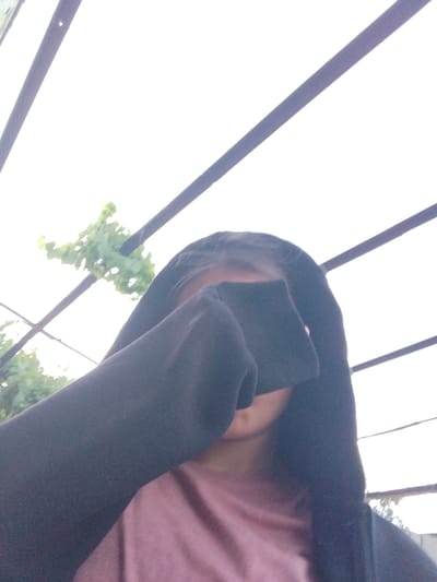 The image shows a person in a close-up shot, covering their eyes with their hand or arm. They are wearing a dark hooded garment, possibly a sweatshirt or jacket, and a pink t-shirt underneath. The background is blurred and appears to be an outdoor setting with green foliage visible to the left, suggesting a daytime scene. Several dark, parallel bars, possibly part of a structure like a pergola or fence, are prominent in the upper and right portions of the frame, set against a bright, overcast sky. The lighting is diffused, indicating either cloudy weather or the subject being in shade. The person's face is mostly obscured, making it difficult to discern any specific facial features or emotions, though a slight smile might be suggested by the visible part of their mouth. The scene appears to be casual and personal, possibly a selfie taken outdoors in Cîrpești, Moldova. There is no visible text in the image.