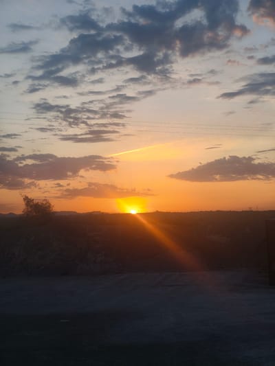 A sunset is captured in Riba-roja de Túria, Spain, with the sun partially obscured by a distant horizon. The sky exhibits a gradient of colors, transitioning from pale blue and pink at the top to vibrant orange and yellow near the setting sun. Wispy clouds are scattered across the sky, some catching the golden light. In the foreground, a dark, silhouetted landscape includes a pile of rocks or debris on the left and a flat, dusty area that appears to be a road or a construction site. Power lines are visible in the upper portion of the image. The overall impression is one of a rural or industrial landscape at dusk.