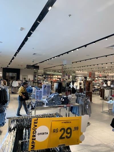 The image captures the interior of a large, well-lit clothing retail store, likely situated within Camará Shopping in Camaragibe, Brazil. The store features a white, dropped ceiling with extensive black track lighting fixtures and recessed illumination. The floor is covered with light-colored, possibly beige, square tiles.
In the immediate foreground, a yellow promotional sign on a clothing rack reads "IMPERDÍVEL OFERTA A PARTIR DE R$ 29,99" (Unmissable Offer Starting from R$ 29.99). The rack below displays numerous dark-colored garments, appearing to be denim.
One individual, an adult female with dark hair, is present in the mid-ground. She is attired in a yellow long-sleeved top and dark trousers, with a dark handbag slung over her left shoulder. She is observed walking towards the left side of the frame, directing her gaze downwards at merchandise presented on a large, rectangular metal display table. This table holds numerous stacks of folded denim jeans and shorts. Smaller display signs featuring models in denim are also on the table.
Beyond the foreground display, the store extends into a spacious area filled with additional clothing racks, display tables, and mannequins showcasing various apparel items, including shirts, blouses, and other casual wear. An overhead black sign with white text points to the left, indicating "CAIXAS / PROVADORES / ATENDIMENTO AO CLIENTE" (Cashiers / Fitting Rooms / Customer Service). A red fire extinguisher is visible mounted on a column in the background. The store appears organized, with neatly arranged merchandise. No other individuals are discernible in direct interaction within the captured scene.