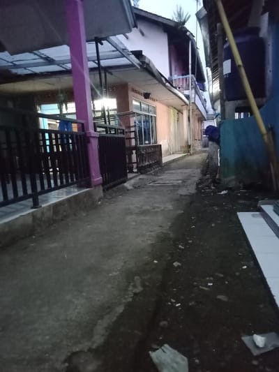 This image depicts a narrow residential alleyway in Ciputri, Indonesia, during low-light conditions consistent with twilight. On the left, a two-story building features a peach-toned ground floor facade with visible wear, a large window reflecting a bright interior light source, and a dark metal vertical-bar fence on a raised concrete base. A prominent magenta-colored support column stands centrally on the left. Above, a corrugated metal awning extends, leading to an upper story with a visible blue balcony railing. Sparse vegetation is observed on the roofline. On the right, a blue-painted wall borders the alley, with a dark blue water tank mounted on a support structure behind a light-colored bamboo pole. The ground comprises a worn, uneven concrete path centrally, flanked on the right by dark, loose soil containing scattered debris, including small white fragments. No individuals are present.