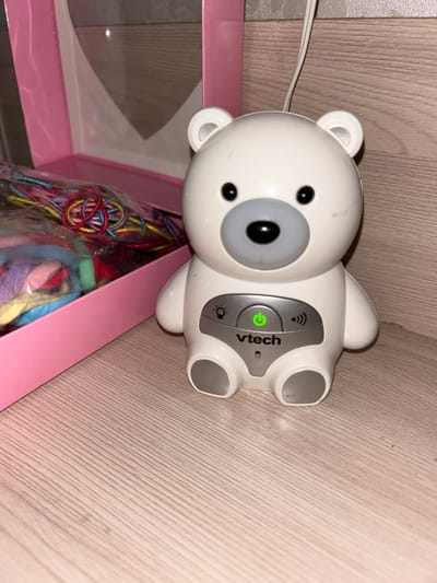 In Primorka, Russia, a white VTech bear-shaped baby monitor is positioned on a light wood-grained surface. The monitor features a grey snout, black eyes, and a green power button illuminated on its chest. To the left of the monitor is a pink box containing a bundle of colorful strings. The background consists of a plain, light-colored wall.