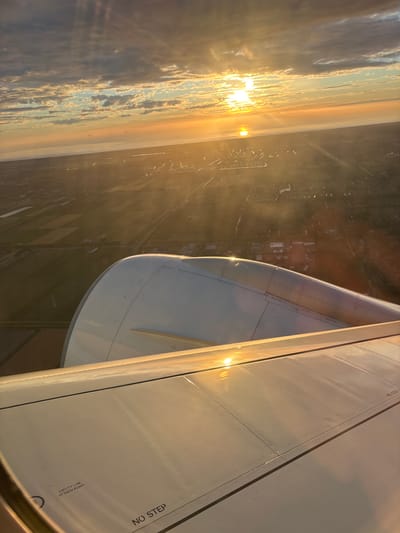 The image captures a view from an airplane window overlooking the city of Nieuw-Vennep, Netherlands, during sunset. The foreground features a portion of the airplane's wing and engine nacelle, reflecting the warm glow of the setting sun. The nacelle is predominantly white with visible seams and rivets, characteristic of aircraft construction. The wing surface is also white and displays markings such as "NO STEP" and "SAFETY LINE ATTACH POINT."
Beyond the aircraft elements, the landscape of Nieuw-Vennep unfolds. The setting sun casts long shadows and creates a golden haze over the scene. The city appears to be a mix of urban development and agricultural land, with visible fields, waterways, and structures. The sky above is dramatic, with a mix of dark and illuminated clouds, and the sun is positioned near the horizon, creating visible light rays. The overall impression is one of a serene aerial perspective of a Dutch landscape at dusk.
