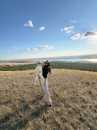In the city of Gornyi Balyklei, Russia, a woman wearing a brown hat, sunglasses, and a black top is holding a young child in her arms. The child is wearing a yellow and white checkered outfit and sunglasses. They are standing on a grassy hill with dry, yellow grass. In the background, there is a body of water, possibly a river or lake, surrounded by green trees and a wide expanse of sky with scattered white clouds. The sun is setting, casting a warm glow over the landscape. The woman appears to be in motion, possibly turning or dancing with the child.