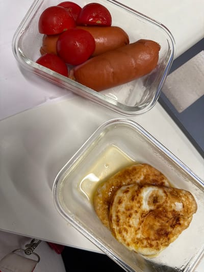 In Kirovsk, Russia, an overhead shot captures two transparent glass containers with food. The upper container holds several whole cherry tomatoes and four sausages. The lower container contains two fried eggs swimming in a light yellow liquid, likely oil or melted butter. The containers sit on a white surface, possibly a table or tray, with a partially visible grey surface to the right.