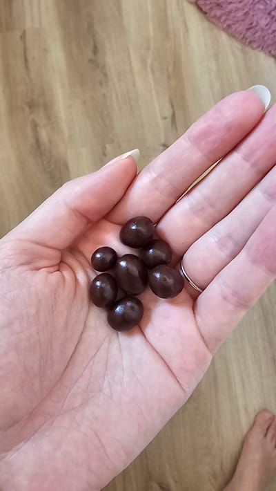 A person's hand is holding a small cluster of dark, oval-shaped candies that appear to be chocolate-covered. The candies are clustered in the palm of the hand, with some resting on the fingers. The skin tone of the hand is light. The background consists of a light brown wooden surface, possibly a floor or table, with a faint pink textured material visible in the upper left corner. A portion of a bare foot is visible in the upper right corner of the image, also resting on the wooden surface. The lighting suggests an indoor setting, possibly during the daytime. There is no visible text in the image.