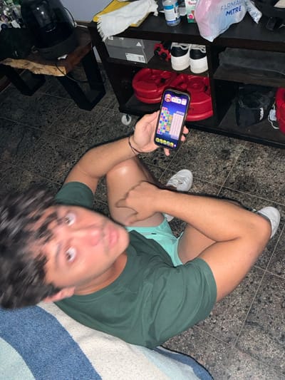 A man with wide eyes and dark curly hair looks up while holding a smartphone displaying a colorful matching game. He wears a green t-shirt and light blue shorts. He sits on a blue and white striped surface. Behind him is a dark shelving unit with shoes, weights, and a bag. The floor is tiled. He points down with his other hand.