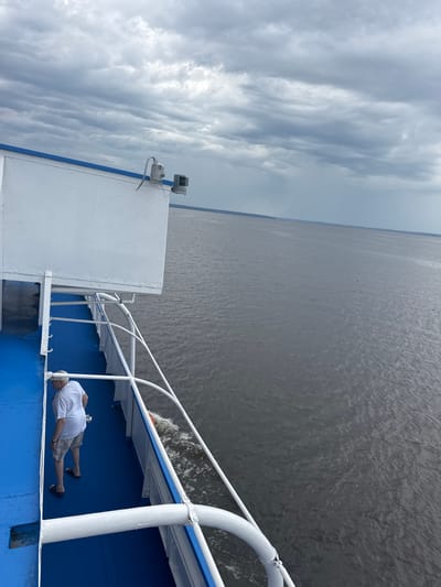 On the deck of a vessel in Ikskoe Ust'e, Russia, a man wearing a white t-shirt and khaki shorts stands near the railing, facing away from the camera and looking out at the water. The deck is painted bright blue and is bordered by a white railing. The vessel is moving, as evidenced by the wake trailing behind it. The sea is a dark gray, and the sky is overcast with heavy clouds. The overall scene suggests a calm but potentially stormy day at sea.