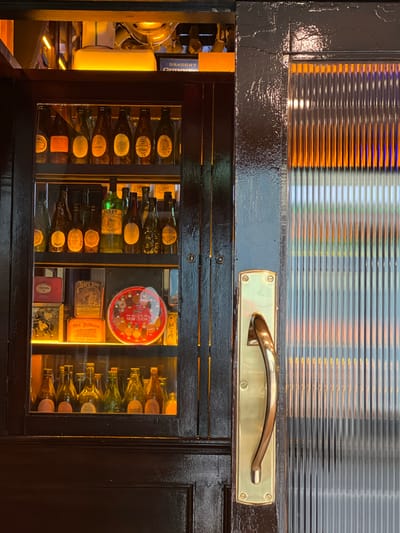 A close-up, eye-level, interior shot captures a dark wooden cabinet with glass doors, revealing a collection of vintage bottles and tins. The cabinet, located in Donegal, Ireland, is illuminated from within by warm yellow lighting. The left side of the cabinet displays several shelves stacked with amber and green glass bottles, some with yellow labels. Below the bottles, there are various vintage tins, including one red tin labeled "CRANOS," a brown tin with a picture of a man and labeled "Sear's Soap," and an orange tin with text that includes "Old Holborn." A round red tin with a picture of biscuits is also visible. The right side of the cabinet features a fluted glass door with a brass handle. Through the fluted glass, blurred orange and blue lights are visible. The overall impression is of a well-preserved collection of historical items in a dimly lit setting.