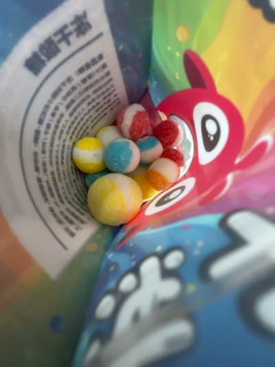 This is a close-up, indoor photograph of a bag of candies. The bag is colorful and features a cartoon character, possibly a rabbit, with a red head and white eyes, set against a background of rainbow colors and stars. Inside the bag, a pile of small, round candies is visible. These candies are multi-colored, with stripes of red and white, blue and white, yellow and white, and orange and white. A portion of a white label with Chinese text is also visible inside the bag. The photograph is taken from a slightly downward angle, looking into the opening of the bag. The overall lighting is bright. There are no people or animals present in the image. The specific location within Yakovlevskoye, Russia, cannot be determined from the visual information provided in the image.