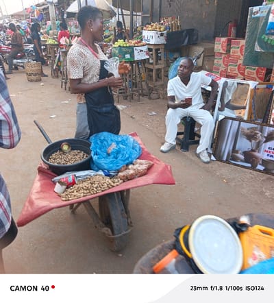 A street scene depicts a woman selling peanuts from a wheelbarrow topped with a red cloth. The wheelbarrow contains a frying pan filled with peanuts, a bag of peanuts, and a blue plastic bag. The woman is holding a plastic bag. In the background, a man dressed in white sits on a stool using a phone. Beside him are boxes, mirrors, and a sign. The scene is bustling with activity in the background, including other vendors and people on motorcycles. The image is captured in warm tones with a shallow depth of field.