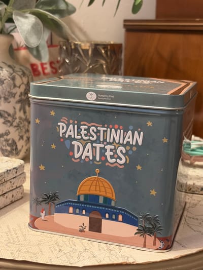 A close-up shot shows a light blue tin of "Palestinian Dates" sitting on a surface covered in a map. The tin features a cartoon illustration of the Dome of the Rock in Jerusalem, complete with palm trees and yellow stars on the blue background. The words "Palestinian Dates" are printed vertically in a colorful, playful font. To the left of the text is the logo for "Humanity First Serving Mankind." The tin sits on top of several small, patterned objects. A plant, partially out of focus, sits to the left of the tin, and a wooden surface is visible in the background. A red sign that reads "BEST" is also partially visible. The city of Reigate and Banstead, United Kingdom cannot be determined from the image.