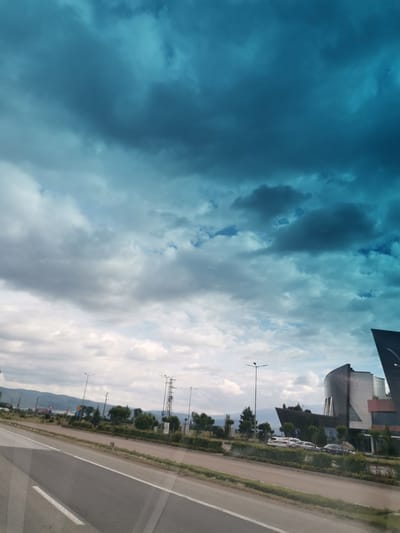The image depicts a road in Krivina, Bulgaria, with a modern building and a dramatic sky. The road in the foreground is paved with asphalt and has white lane markings. To the right of the road, there is a modern building with a distinctive, curved, dark-colored facade and glass elements. Several cars are parked in front of the building. The surrounding area includes green landscaping and trees. In the background, there are hills and mountains. The sky is filled with dark, brooding clouds, suggesting an impending storm or a dramatic weather event, with lighter clouds and a hint of blue sky peeking through. The overall impression is one of an urban or semi-urban environment under a powerful sky.