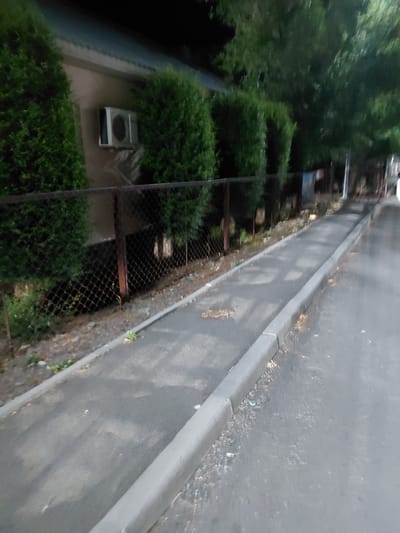 The image depicts an outdoor scene, possibly a residential area, with a focus on a road and adjacent greenery. A paved road runs along the right side of the frame, bordered by a concrete curb. The road surface is a dark gray asphalt, with some scattered debris and fallen leaves. The curb is a lighter shade of gray and appears to be well-maintained.
To the left of the road, there is a strip of vegetation consisting of manicured bushes and trees, creating a green buffer. Further to the left, a chain-link fence separates the vegetation from what appears to be a lower-lying area, possibly a drainage ditch or a walkway. Through the fence, more dense greenery is visible, along with some dark, shadowy areas.
In the lower left quadrant of the image, a silver appliance, possibly an air conditioning unit or a ventilation component, is visible surrounded by bushes. Some rusted metal structures and possibly wooden planks are also discernible in the middle ground, integrated with the fence line. The overall impression is one of an urban or suburban setting with an emphasis on landscaping and infrastructure.
The lighting suggests it could be late afternoon or early evening, with soft shadows cast across the scene. The weather appears to be fair, with no signs of rain or extreme conditions. No people are visible in the frame. The image is taken from a slightly elevated perspective, looking down and across the scene. The location context provided is Ақши, Kazakhstan. No visible text is present in the image.