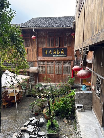 The image displays an exterior view of a traditional Chinese courtyard in Gongtan, China. A multi-story wooden building, identified as "宜安居客栈" (Yi An Guesthouse) by a prominent black sign with gold characters and a phone number (18996982030), dominates the background. This guesthouse features multiple latticed windows across its levels and a dark, tiled roof with a visible metal downspout. Red lanterns are suspended from its eaves, and a woven basket hangs from its second story. To the right, a second multi-story wooden structure with traditional architectural elements and a red lantern is partially visible. The paved courtyard ground appears wet. On the left, a white canvas tent covers an outdoor seating area containing multiple wooden tables and chairs. A pile of hollow grey concrete blocks is stacked in the foreground-left. Various potted plants and small garden beds are arranged throughout the courtyard, displaying diverse foliage. A small, blue plastic stool is positioned near the right building. No individuals or specific actions are present within the scene. The sky is overcast.