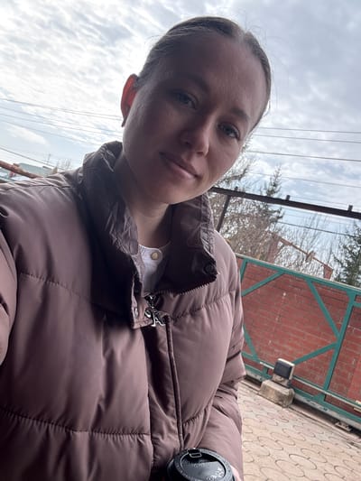 An individual, appearing female, with fair skin, light-colored eyes, and light hair pulled back, is depicted in a selfie taken outdoors in Gamovo, Russia. They are wearing a brown/mauve puffer jacket over a white collared top with a visible golden button. A black disposable beverage cup with a lid is held in the lower right of the frame. The subject is looking directly at the camera. The background features an overcast sky with scattered clouds. Visible behind the subject are several utility wires, bare deciduous trees, and coniferous trees. To the right, a red brick wall is partially visible, fronted by a green metal fence or gate structure. The ground in the lower right consists of patterned paving stones. A concrete block supporting a dark, rectangular object is present on the paved ground near the fence.