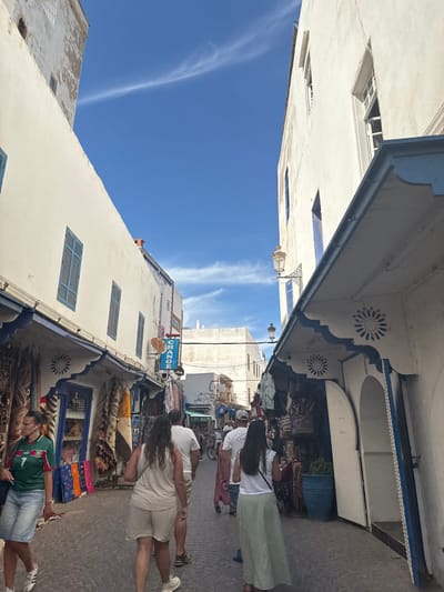 This is an eye-level, medium shot of a narrow, cobblestone street in Essaouira, Morocco, lined with shops on both sides. The buildings are whitewashed with blue trim and accents on the awnings and windows. Sunlight illuminates the scene, casting shadows on the ground.
In the foreground, a woman wearing a green and red soccer jersey with the number "7" and denim shorts walks to the left, looking to her left. To her right, a person wearing a light-colored t-shirt and shorts walks away from the camera, followed by another person in a white top and long, pale green skirt. Several other people are visible walking further down the street, some with bicycles.
The shops display a variety of goods, including textiles and rugs hanging outside. A blue sign that reads "CHANGE" is visible on the right side of the street. The sky is a vibrant blue with a few wispy white clouds. Traditional Moroccan architectural elements, such as decorative windows and arched doorways, are present. The overall atmosphere suggests a bustling marketplace or a busy tourist area.