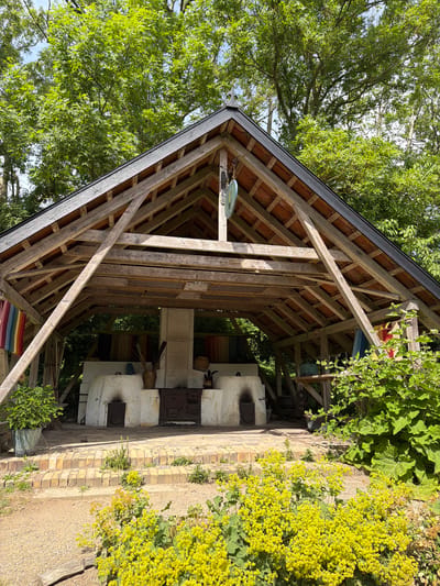 In Lejre, Denmark, this image shows an open-air structure with a wooden roof supported by timber beams. The structure shelters a large, white, oven-like structure with two openings in front. A dark-colored stove or oven is positioned in the center, in front of the white structure. Various items, including pots and what appear to be bundles of sticks, are visible around the ovens. Colorful textiles, possibly tapestries or cloths, hang from the sides of the structure. The area in front of the structure is paved with bricks and leads to a dirt path. Lush green foliage and bright yellow flowers surround the scene, with large trees forming a backdrop. The sky is visible through the leaves of the trees, suggesting it is daytime. There are no people or animals visible in the image.