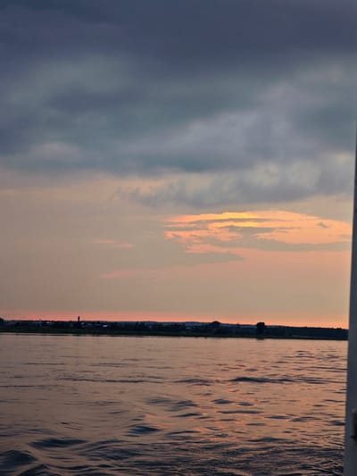 The image depicts a panoramic view of a wide river at dusk, with a distant treeline and settlement along the far bank. The sky is filled with dramatic, layered clouds in shades of gray, deep blue, and soft orange, suggesting a sunset or sunrise. The water's surface is gently rippled, reflecting the muted colors of the sky. The settlement on the far bank appears to be a small town or village, with scattered buildings and trees visible against the horizon. A prominent structure, possibly a tower or antenna, stands out against the skyline. The overall atmosphere is serene and natural, with a touch of dramatic beauty due to the cloud formations and the reflected light on the water. The scene is likely captured from a boat or the shore of the river, looking across towards the opposite bank.
