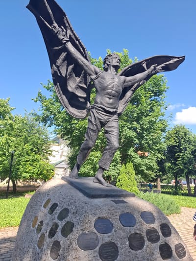 A bronze statue of a winged man is displayed outdoors against a bright blue sky, surrounded by lush green trees. The statue depicts a muscular, semi-nude figure with outstretched arms and large, dark wings. The man's face shows a strained expression, and his body is contorted as if in mid-flight or struggle. He is clad in draped garments that resemble a cape or large fabric billowing behind him.
The statue stands on a rough, light-colored stone base embedded with numerous circular bronze plaques, each bearing visible text. These plaques appear to be commemorative, possibly listing names or significant dates. The base is situated in a park-like setting with neatly mown grass and a paved walkway visible in the background. Sunlight casts shadows, suggesting it is daytime, likely morning or afternoon. The overall mood of the statue is one of drama and perhaps heroism or sacrifice. The location is identified as Kungur, Russia.