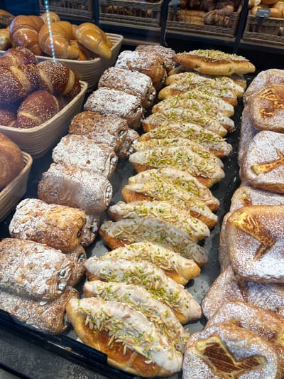 This is a close-up, eye-level, over-the-shoulder shot of a display case filled with a variety of pastries. The pastries are arranged in neat rows and baskets on a black surface. The primary subjects are croissants and other baked goods. Some croissants are plain and golden brown, others are topped with powdered sugar, sliced almonds, and pistachios, and a light glaze. There are also darker, chocolate-filled pastries dusted with powdered sugar and almonds. On the right side of the display, there are pastries with a cream filling and a decorative lattice pattern, also dusted with powdered sugar. The lighting is bright, creating reflections on the glass display case. The background shows additional baskets of pastries and some signage, which appears to be in German. The scene is likely a bakery or a patisserie. The presence of German signage and the context of a bakery setting suggest this image was taken in Esslingen am Neckar, Germany. There are no people visible in the image.