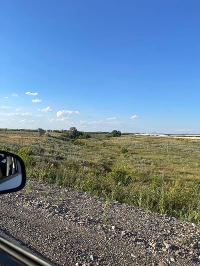 A rural landscape is captured from the perspective of a vehicle's side mirror, looking out at a vast, grassy field under a bright blue sky dotted with scattered white clouds. The foreground is dominated by a rough, gravelly roadside. In the middle ground, the field rolls gently, covered in dry grass and sparse green bushes. Several wooden posts with orange tops are visible in the distance, suggesting some form of infrastructure or markers. In the far background, the land continues to undulate, meeting the horizon with a hint of distant hills or mountains under the expansive sky. The scene appears to be located in Praporştşikovo, Kazakhstan, a region known for its expansive steppes and rural beauty. There are no people or animals visible in the image.