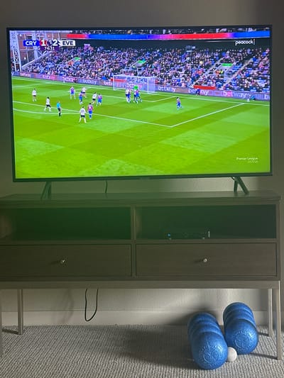 The image shows a television displaying a soccer game between Crystal Palace (CRY) and Everton (EVE), with the score 1-2 in favor of Everton at the 74:31 mark. The game is being broadcasted on Peacock and is part of the Premier League 2025/26 season. The television is mounted on the wall next to a tall, dark grey cabinet with two doors. To the right of the cabinet, there are two blue textured rollers with a white object sandwiched between them. The wall is covered with a grey carpet. The setting appears to be indoors, likely in a private residence, with the geographical location in the city of Clifton, United States of America.
