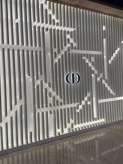 The image shows a wall with a textured surface made of vertical slats of varying lengths and depths, creating a geometric pattern. A illuminated logo, appearing to be "CD", is centrally placed on the wall. The lighting from behind the slats casts shadows, highlighting the three-dimensional effect of the design. The wall is situated in an indoor environment with a reflective floor that mirrors the pattern of the wall and the logo. There is a wooden ceiling visible at the top of the image. The scene does not contain any people or depict any actions or interactions. The provided context suggests the location is Xuanwu District, China, but this is not visually evident in the image itself.
